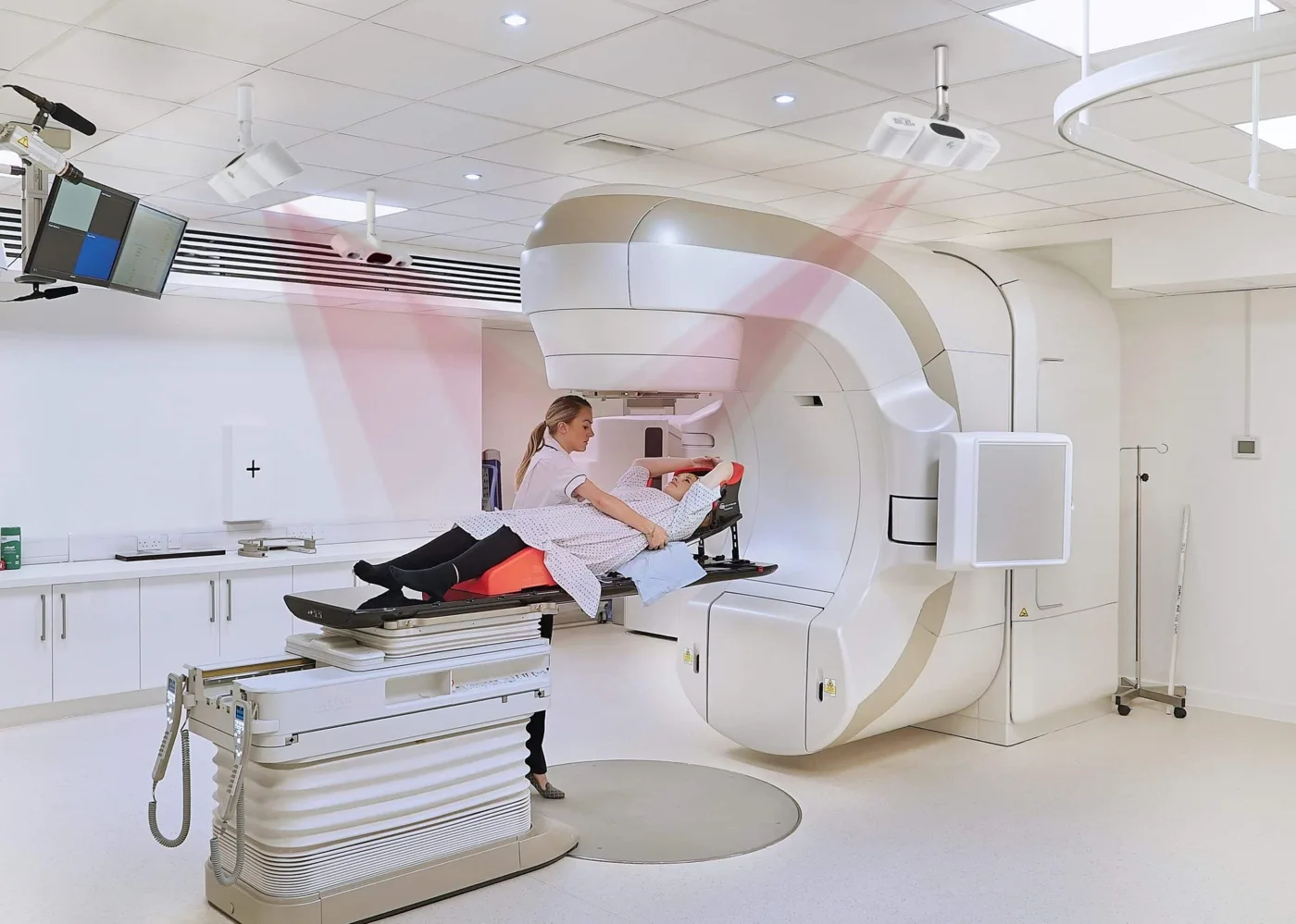 A radiographer positions a patient on the couch of a linear accelerator while ceiling-mounted cameras project red beams onto the patient's body